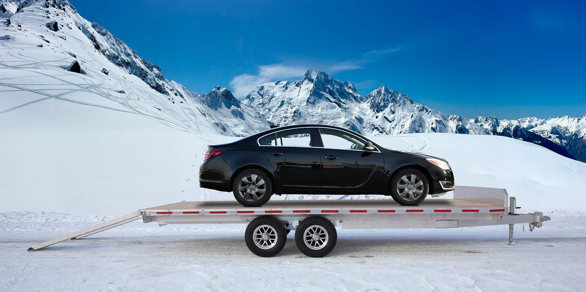 Black sedan loaded on a silver aluminum car hauler trailer against a snowy mountain backdrop.