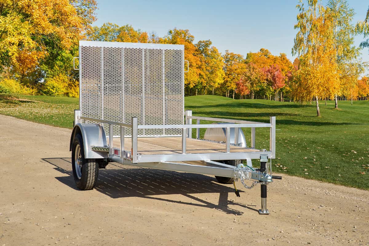 A small silver aluminum utility trailer with mesh rear gate and wood floor parked on a gravel path surrounded by autumn trees.