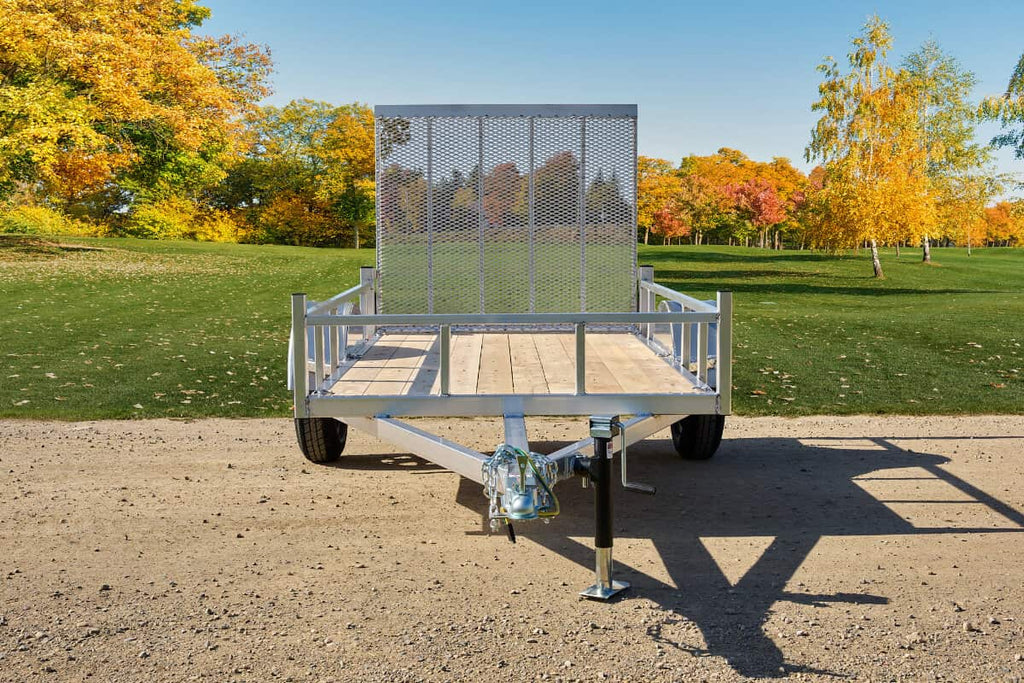 Galvanized single-axle utility trailer with wood floor, mesh front ramp, and tubular side rails parked on a gravel path.