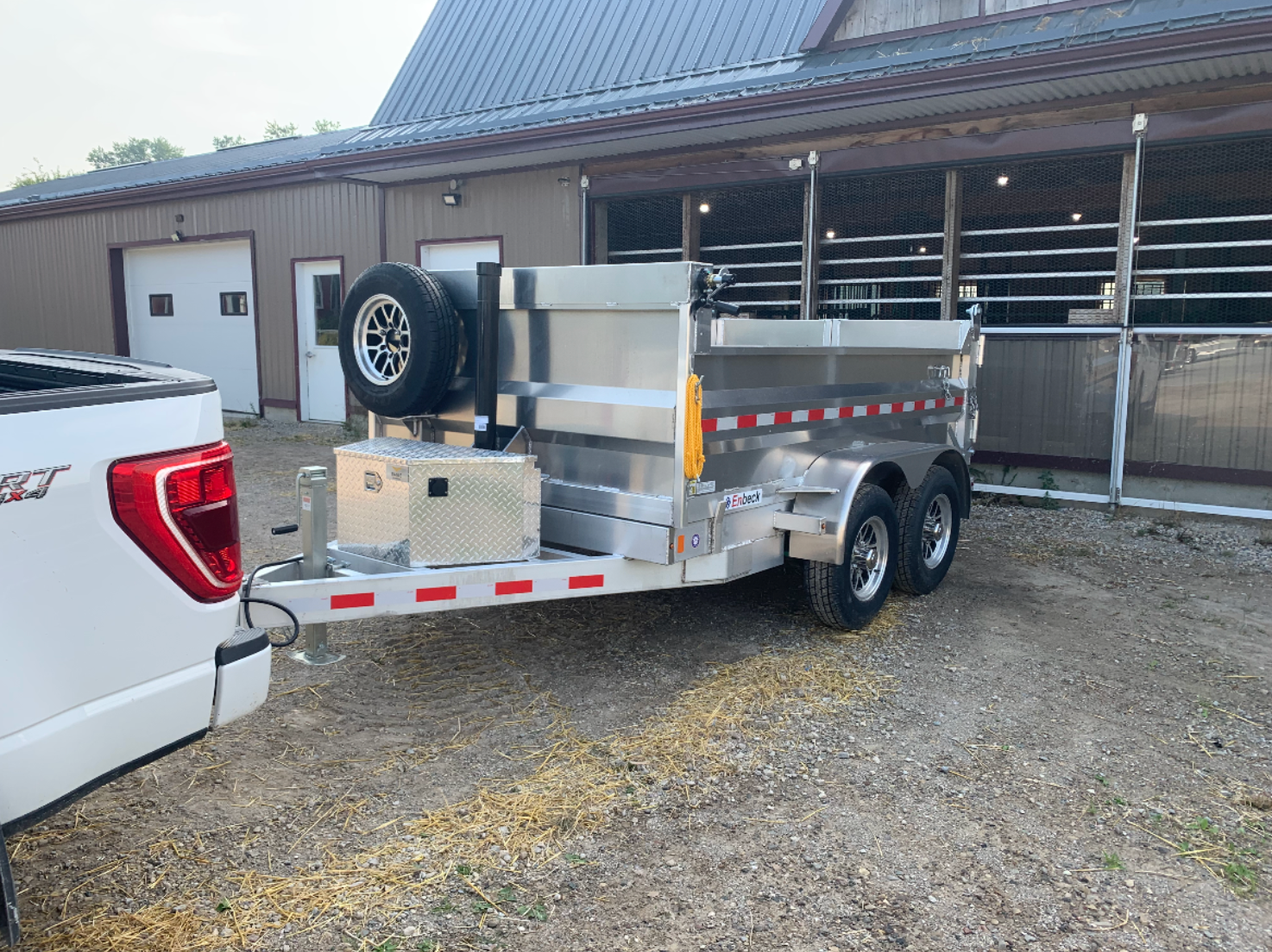 Silver Enbeck aluminum dump trailer with dual axles and spare tire hitched to a white pickup truck in front of a barn.