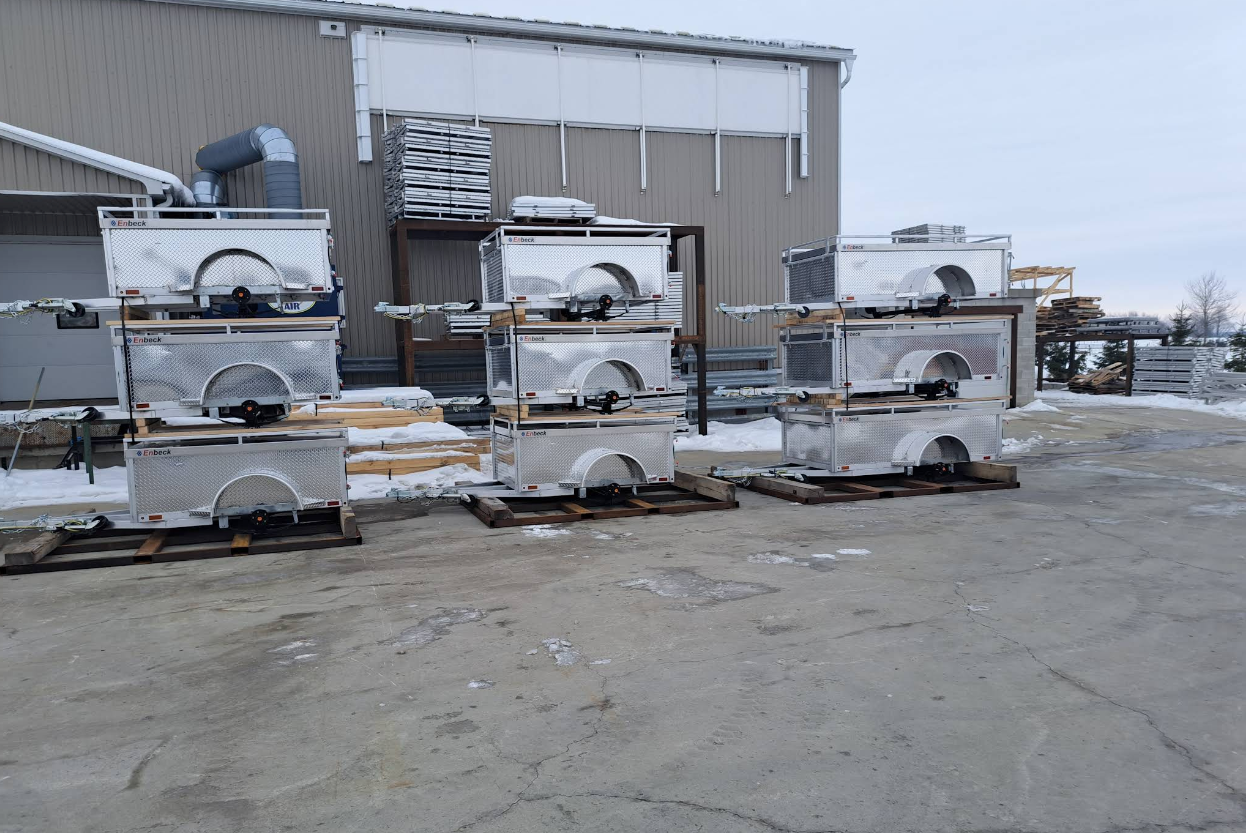 Stacked silver Enbeck aluminum utility trailers on pallets outside an industrial building in snowy conditions.