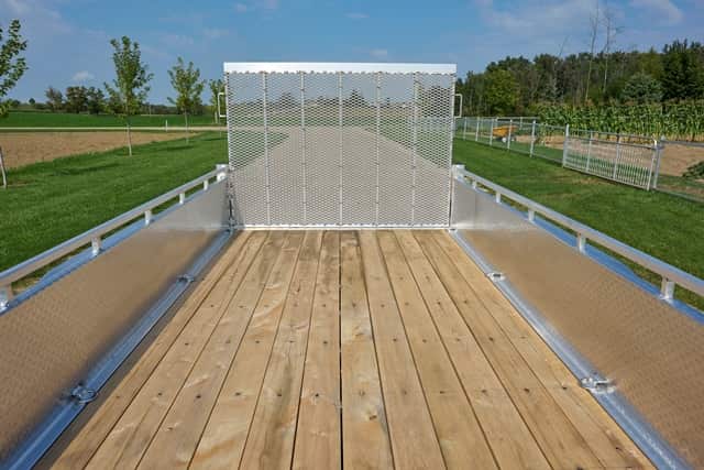 Rear view of an open flatbed utility trailer with a wooden deck, aluminum side rails, and a mesh ramp gate, parked outdoors beside a grassy field.