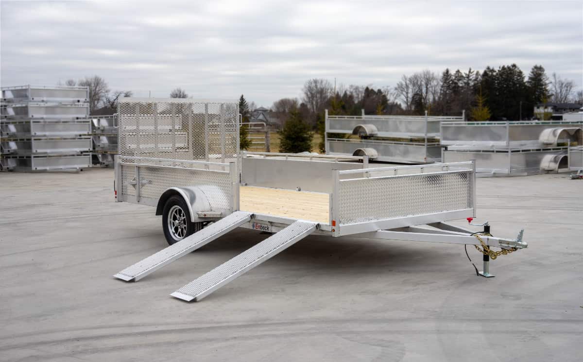 Silver aluminum utility trailer with mesh sides, lowered rear ramp, and wood floor, parked on a lot with other trailers in the background.