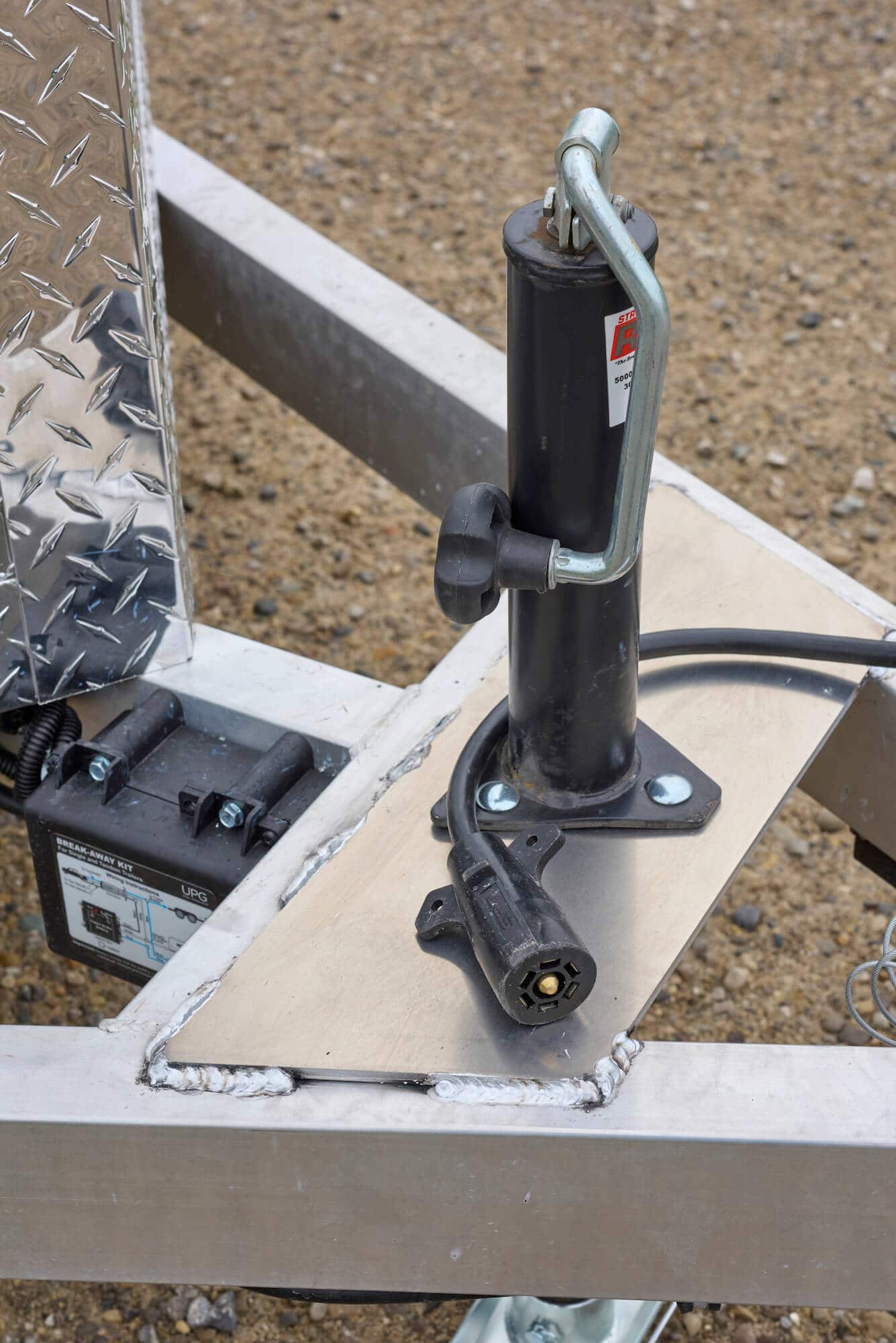 Close-up of a trailer tongue with a black manual jack, crank handle, and electrical connector plug.