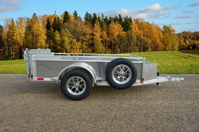 Silver aluminum Enbeck utility trailer with diamond-plate sides on a gravel road, autumn trees in background.