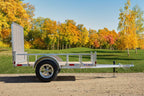 Side view of a silver Enbeck aluminum utility trailer with wood floor and mesh front gate parked on a gravel path amid fall foliage.