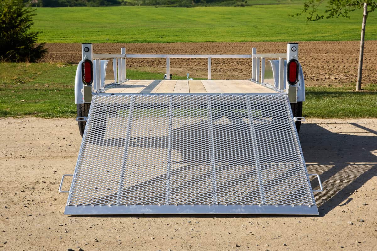 Silver utility trailer with lowered mesh rear ramp gate, viewed from behind on a gravel path in a green field.