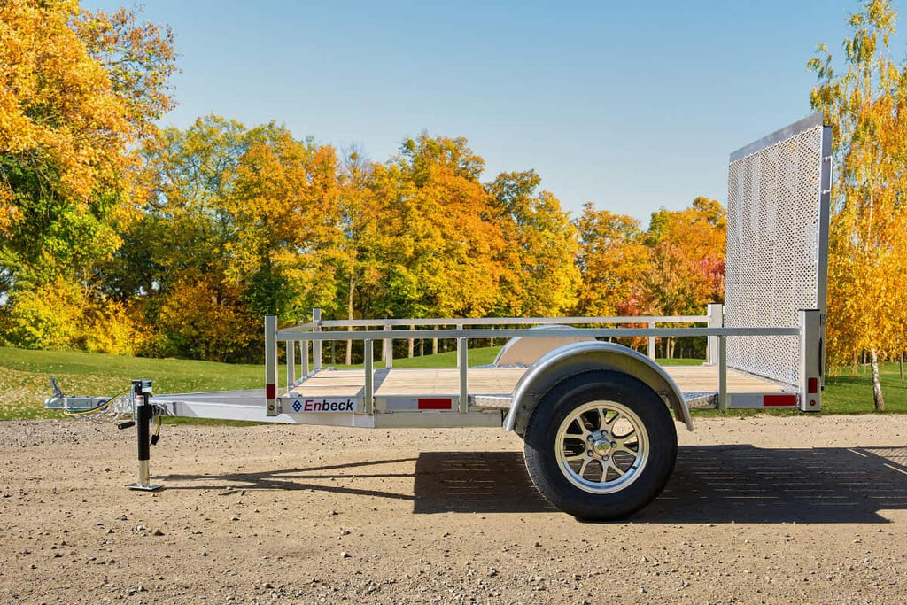 Side view of an Enbeck single-axle galvanized utility trailer with wood floor, mesh rear ramp, and aluminum wheels parked on gravel.