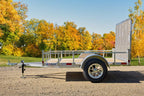 Side view of an Enbeck single-axle galvanized utility trailer with wood floor, mesh rear ramp, and aluminum wheels parked on gravel.