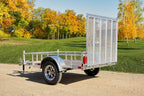 Rear view of a silver Enbeck aluminum utility trailer with mesh tailgate and wood floor on a gravel path amid autumn trees.