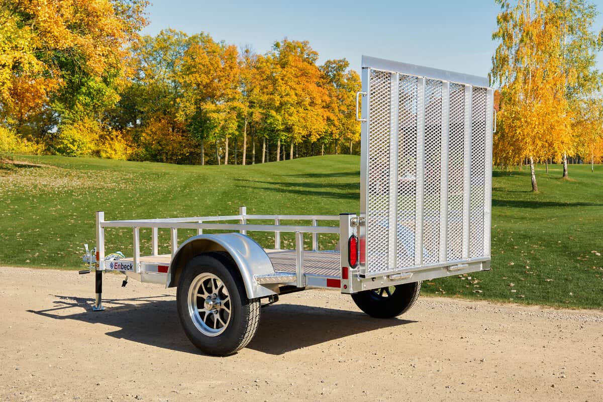 Rear view of a silver Enbeck aluminum utility trailer with mesh tailgate and wood floor on a gravel path amid autumn trees.