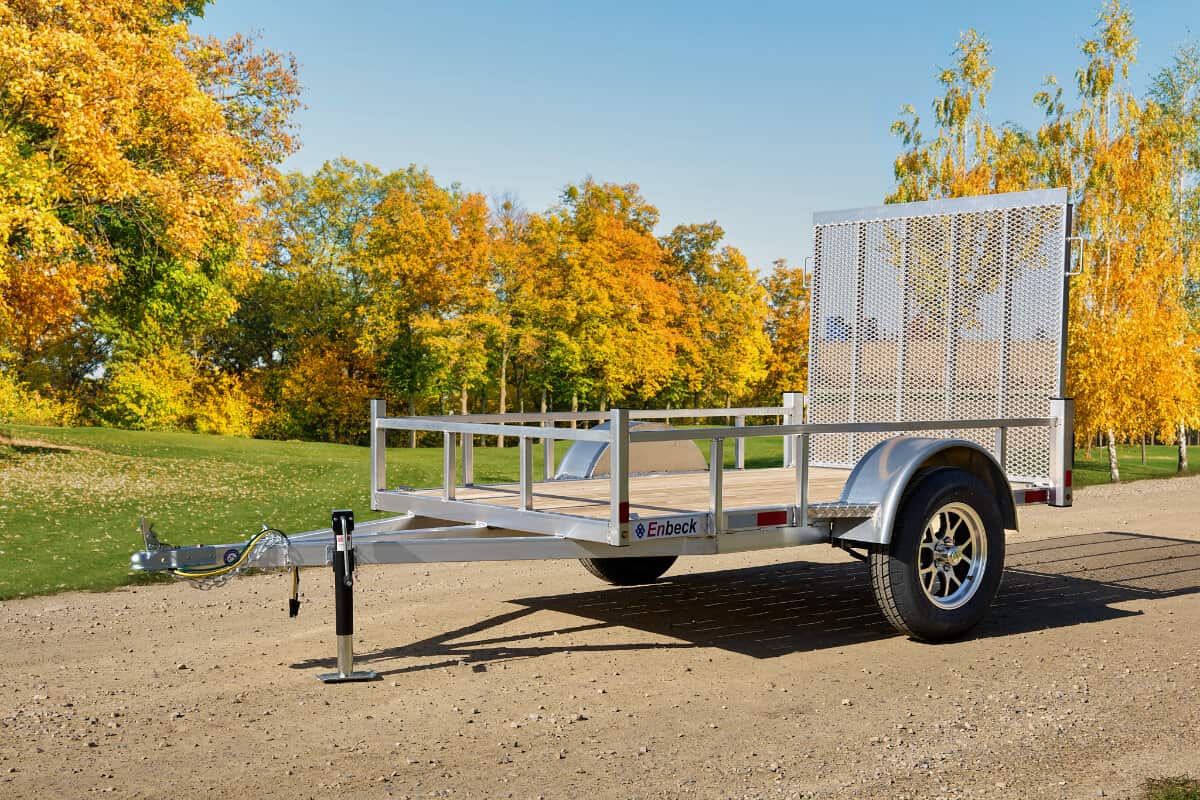 Enbeck single-axle utility trailer with wood floor, mesh side rails, and front ramp parked on gravel amid autumn trees.