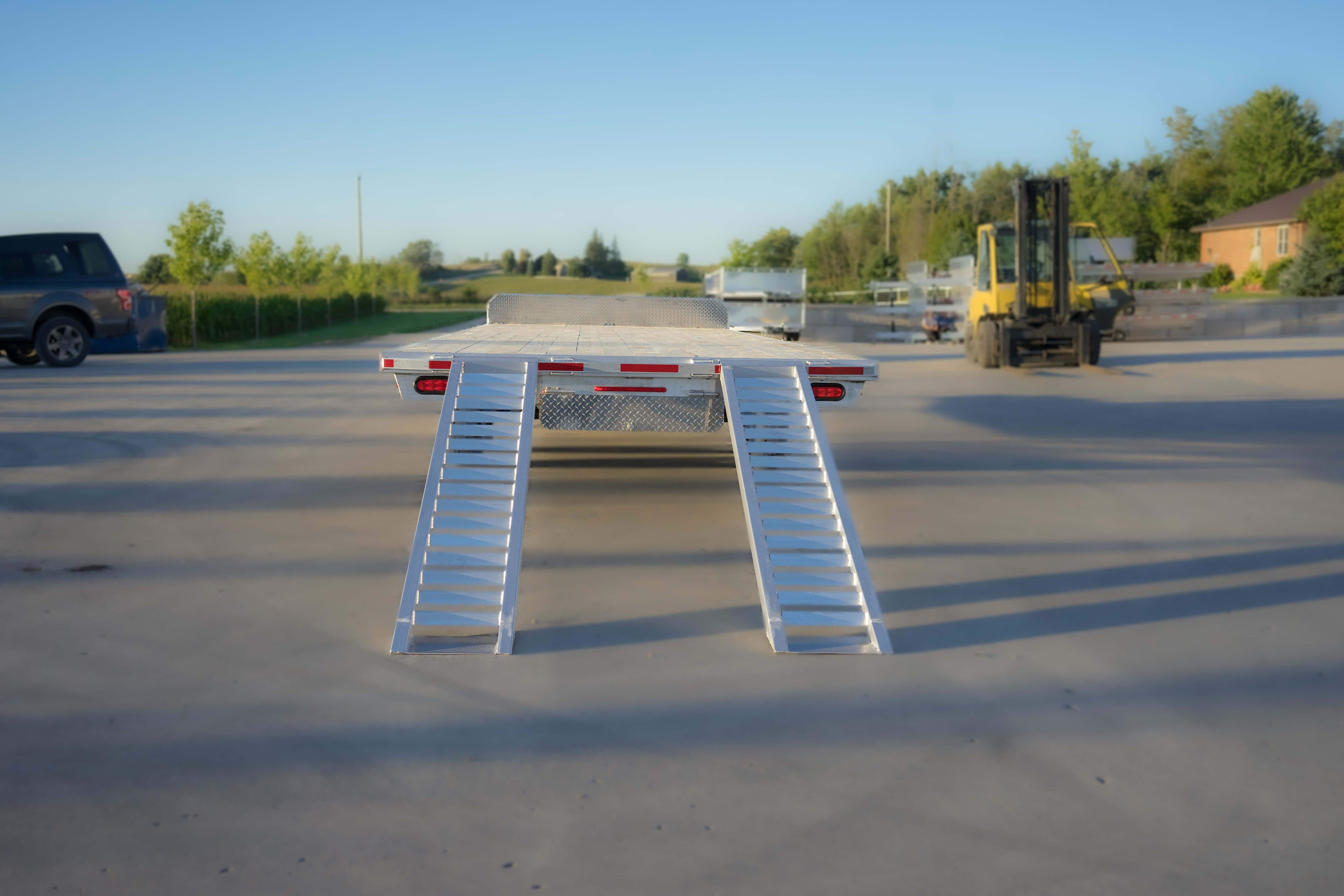 Rear view of an aluminum flatbed trailer with lowered bi-fold loading ramps on a concrete lot.
